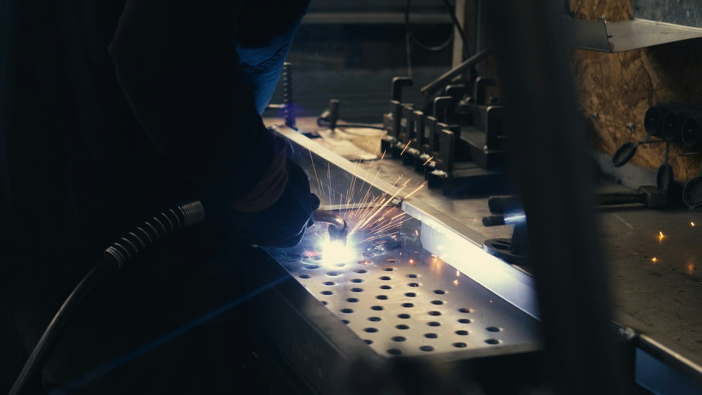 Trabalhador solda uma peça de metal em uma fábrica, com faíscas e equipamento de proteção